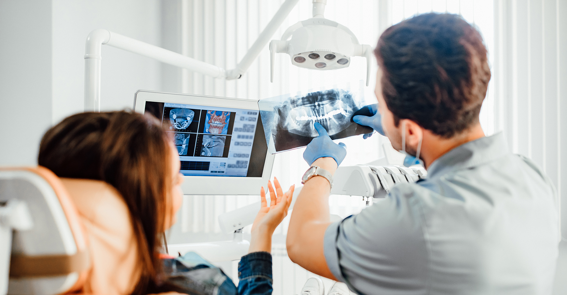 A dental professional showing a patient an x-ray of their teeth on a screen, with both individuals wearing masks, set against a backdrop of a dental office.